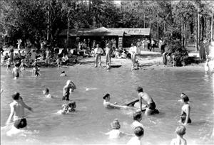 White people of all ages and genders in the water and on the beach. Beyond the beach is forest. There is a large stone structure on the beach with the words men and women carved at either side. 